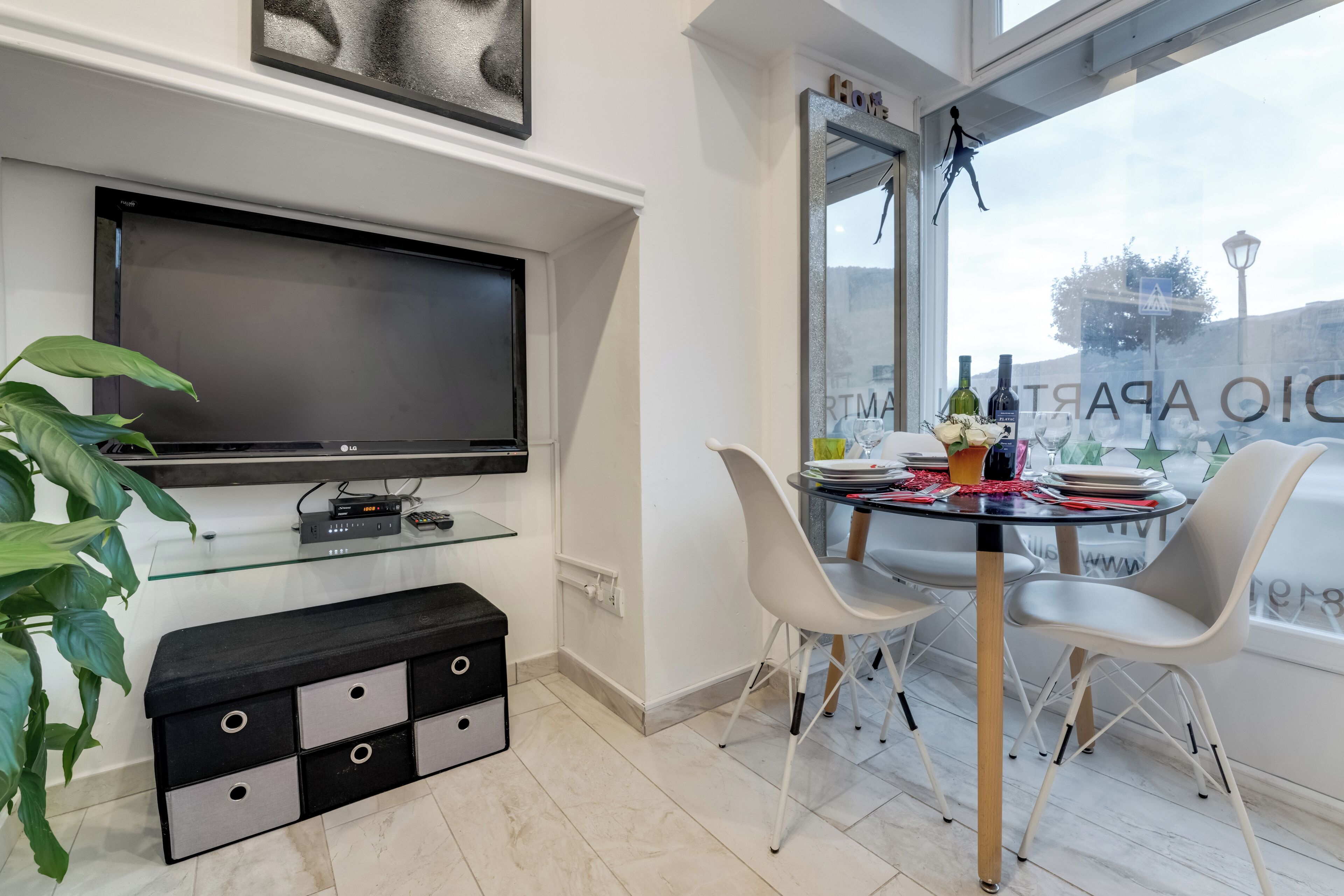 Ett modernt vardagsrum med en platt-TV, ett matbord med glasskiva, vita stolar och en krukväxt.