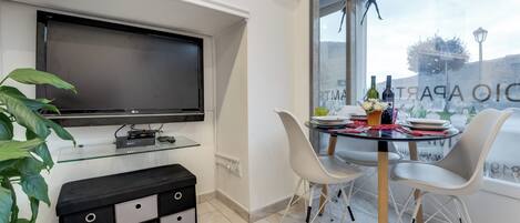 Ett modernt vardagsrum med en platt-TV, ett matbord med glasskiva, vita stolar och en krukväxt.