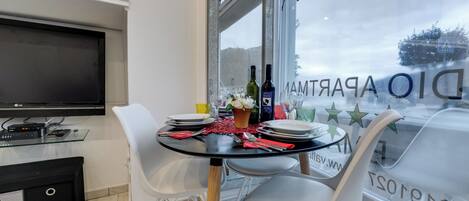 A dining area with a glass table, white chairs, a TV, and a window with a view of the street.