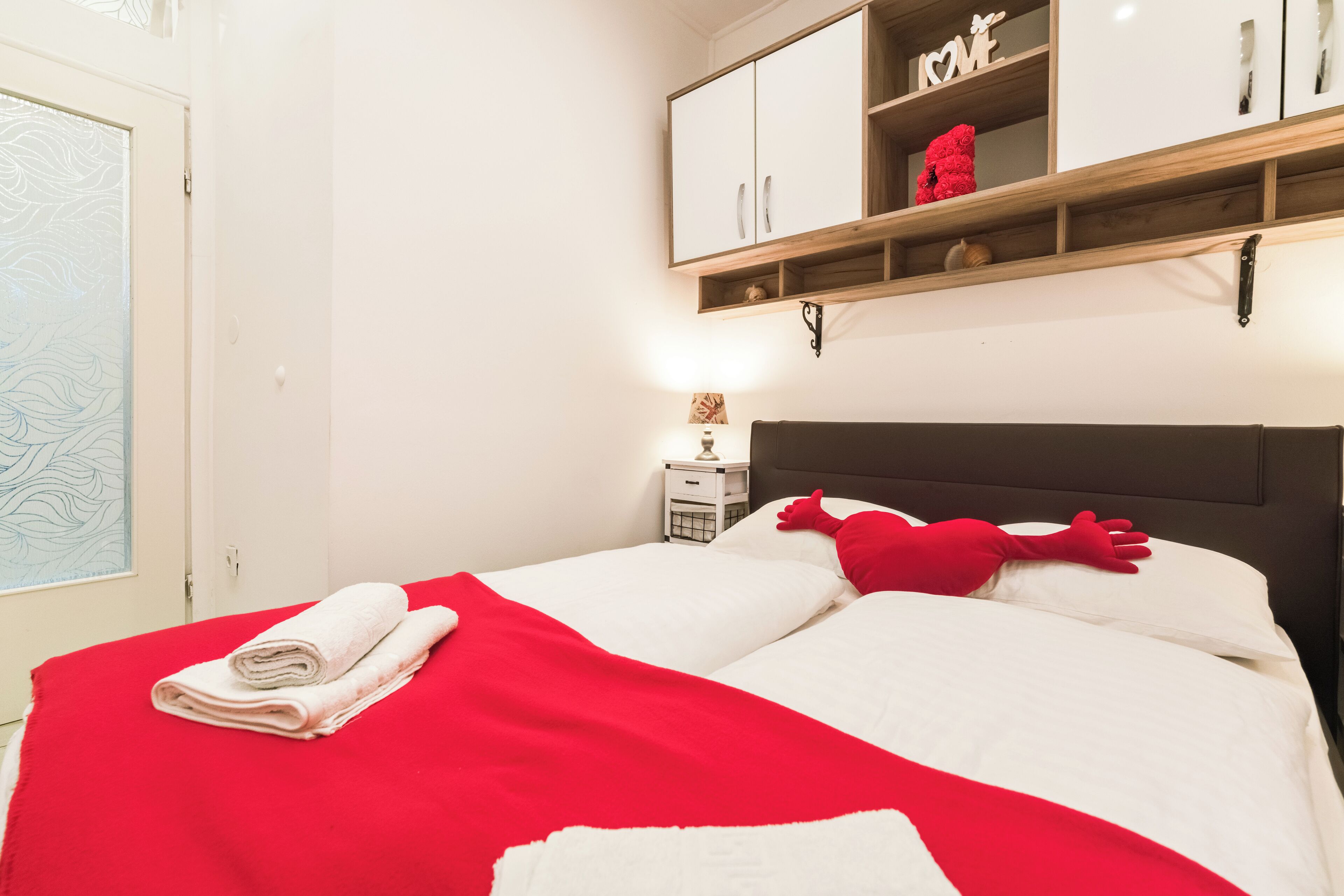 Ein ordentlich bezogenes Bett mit roter Decke und Kissen, ein weißer Nachttisch mit Lampe und ein Holzregal über dem Bett mit Dekoration.