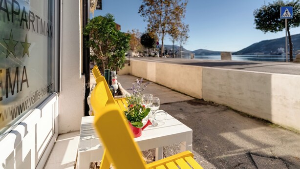 Ein Tisch mit Stühlen vor einem Gebäude, ein Fenster mit einem Schild und Blick auf das Meer und die Hügel.