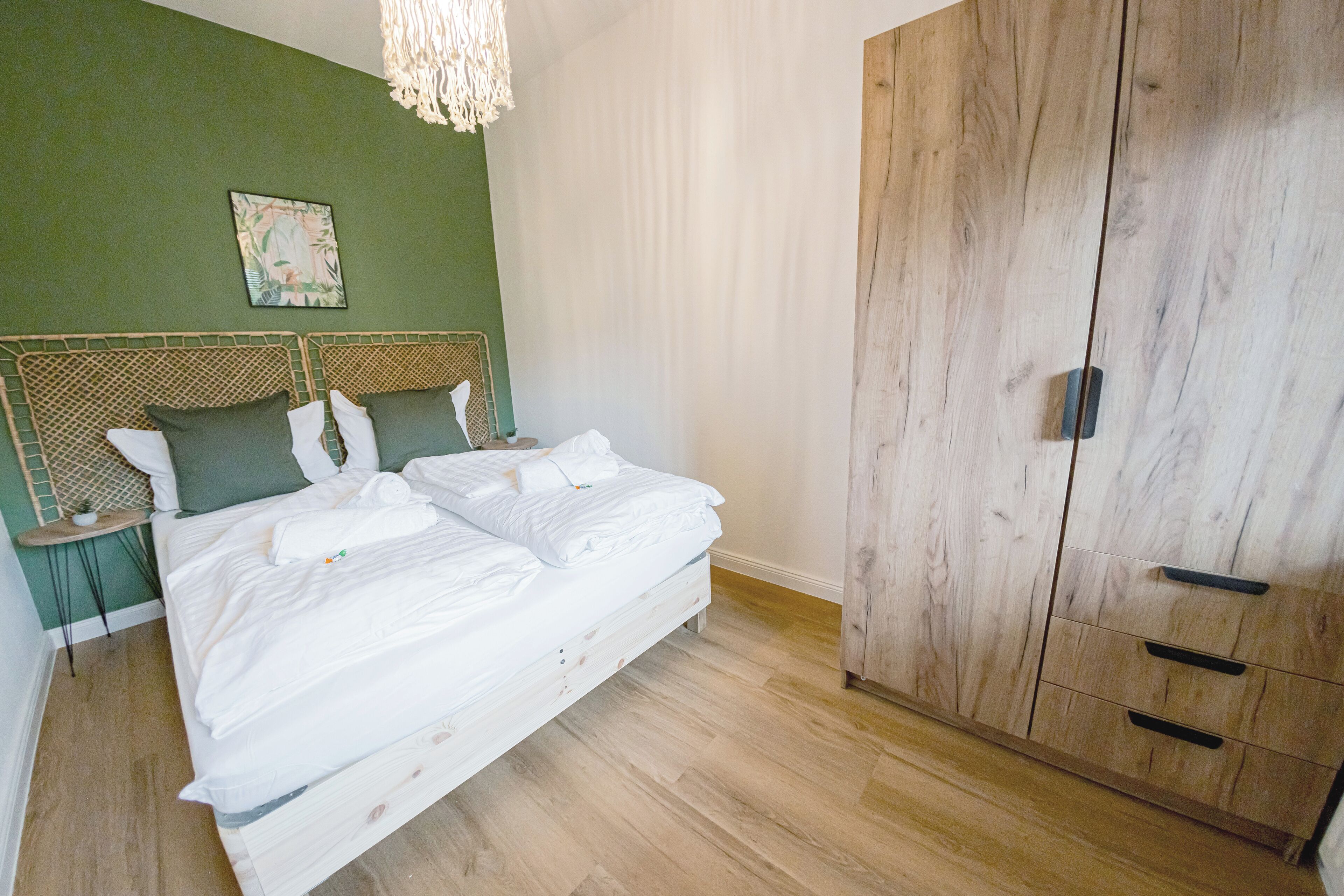 Ein ordentlich bezogenes Bett mit weißen Bettwäsche, ein Holfachschrank mit mehreren Schubladen und eine grüne Wand mit einem gerahmten Bild.
