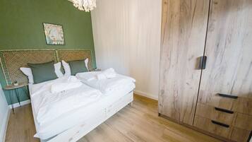 Ein ordentlich bezogenes Bett mit weißen Bettwäsche, ein Holfachschrank mit mehreren Schubladen und eine grüne Wand mit einem gerahmten Bild.