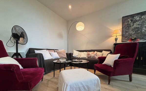 Un salon comprenant un canapé, deux fauteuils, une table basse, une lampe sur pied, un ventilateur et un tableau accroché au mur.