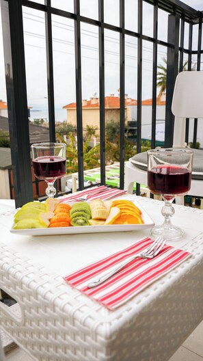 A table with a plate of fruit and two glasses of red wine, set against a window with a view of buildings and palm trees. - Anzede Konaklama Çeşme (Çesme)
