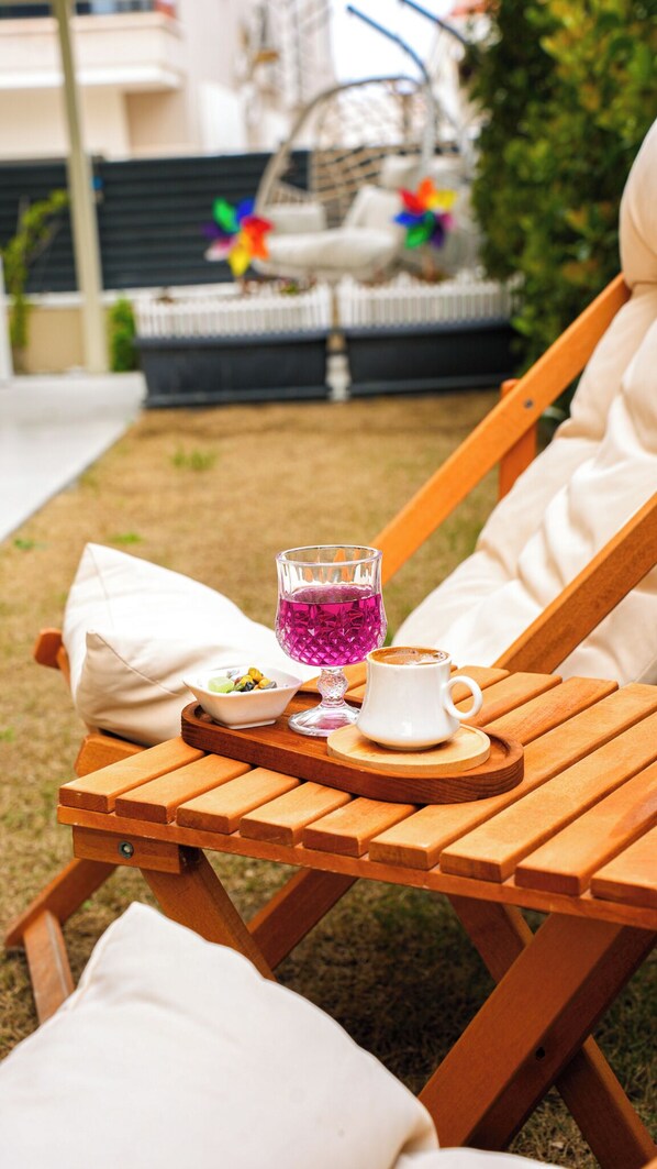 A wooden lounge chair with a tray table holding a glass of purple drink, a white mug, and a small bowl of snacks.
