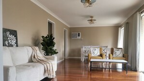 Ein großzügiger Wohnraum mit einem weißen Sofa, einem Couchtisch und einem gelben Sessel. Eine große Pflanze und ein Gemälde sind an der Wand zu sehen.