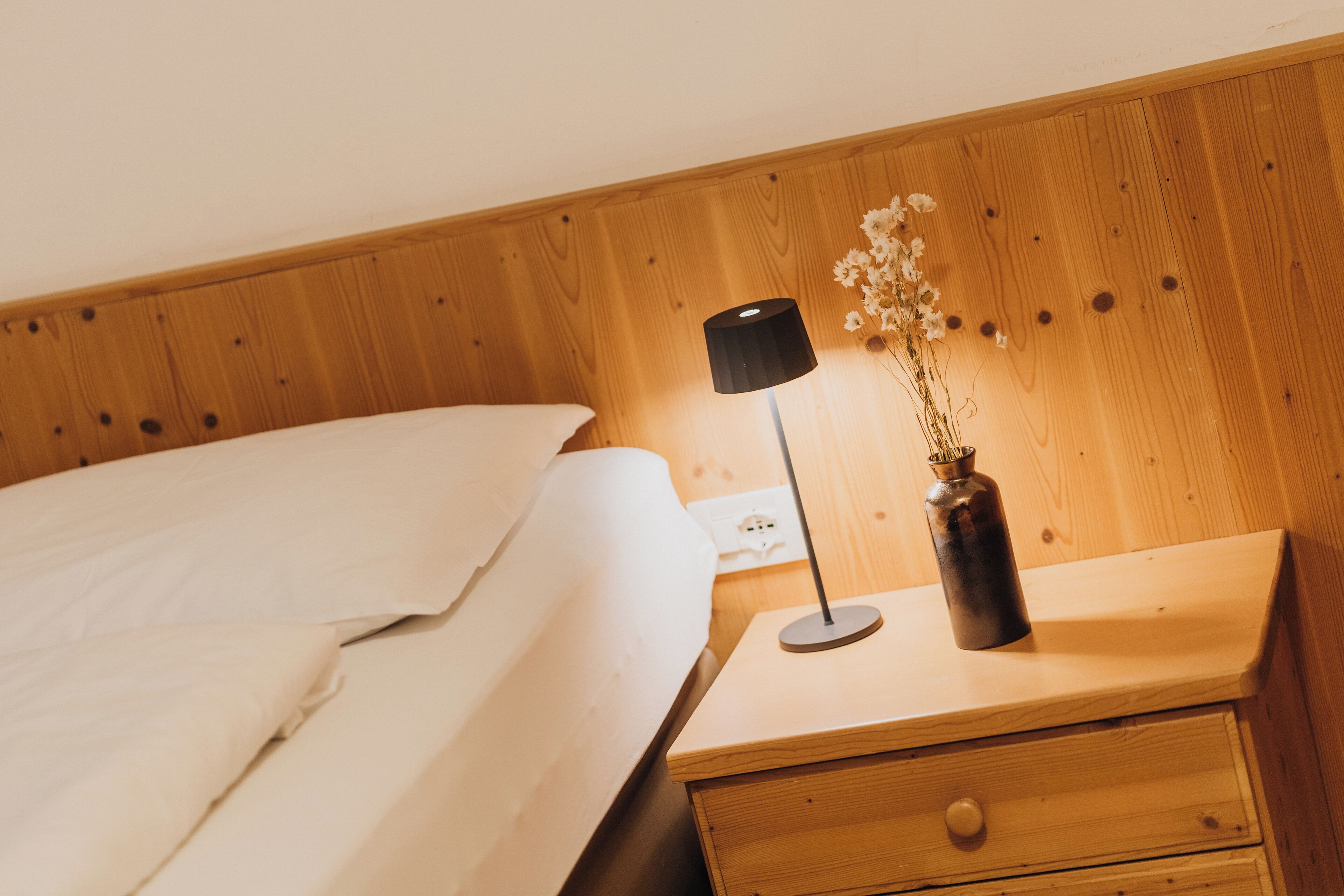 A neatly made bed with white linens, a bedside table with a lamp and a vase of flowers, and a wooden headboard.