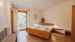 A bedroom with a wooden bed, a bedside table with a lamp, a window with curtains, and a plant on the nightstand. - Appartements Elke (Lagundo)