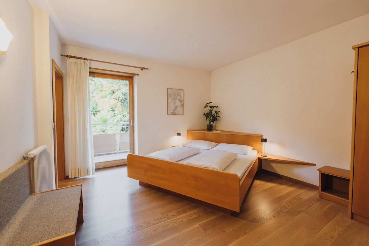 Una camera da letto con un letto in legno, un comodino con una lampada, una finestra con tende e una pianta sul tavolino di fianco al letto.