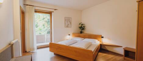 A bedroom with a wooden bed, a bedside table with a lamp, a window with curtains, and a plant on the nightstand.