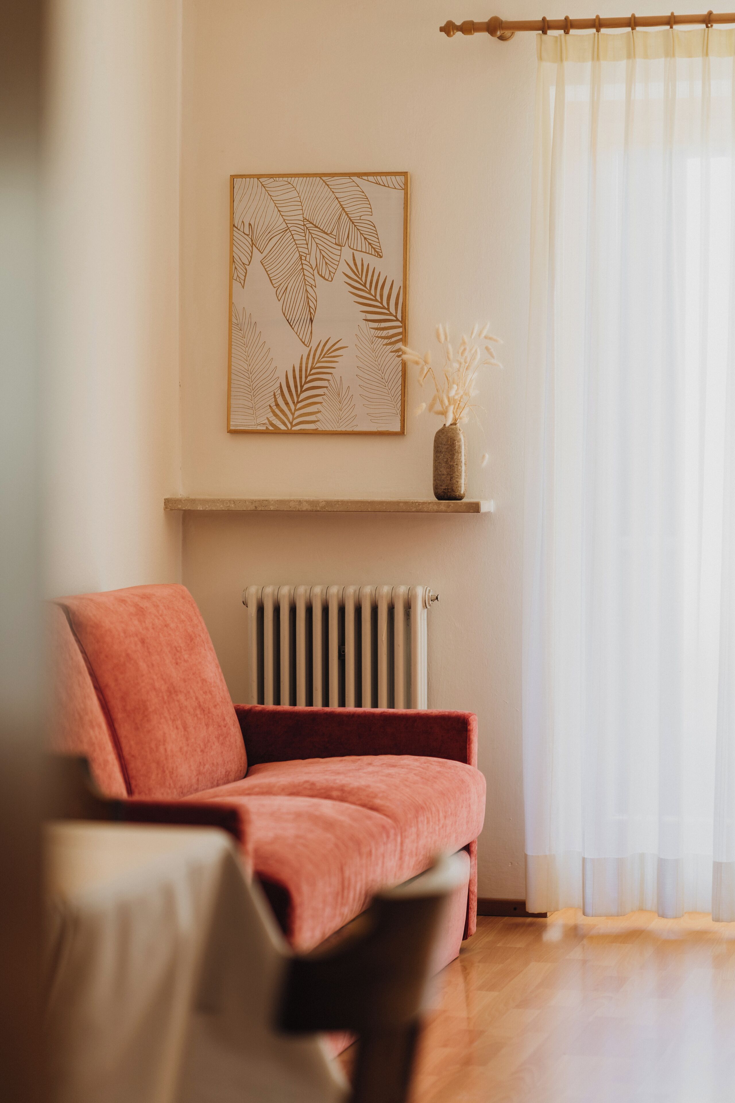 A living room with a red sofa, a radiator, a framed leaf pattern on the wall, and a window with sheer curtains.