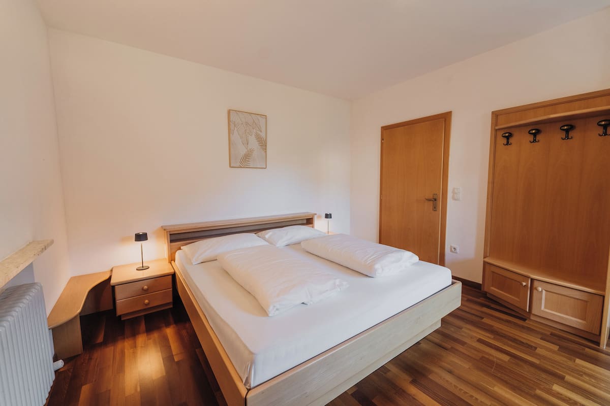 Un letto rifatto con lenzuola bianche, un comodino con una lampada, un armadio in legno e un termosifone a pavimento.