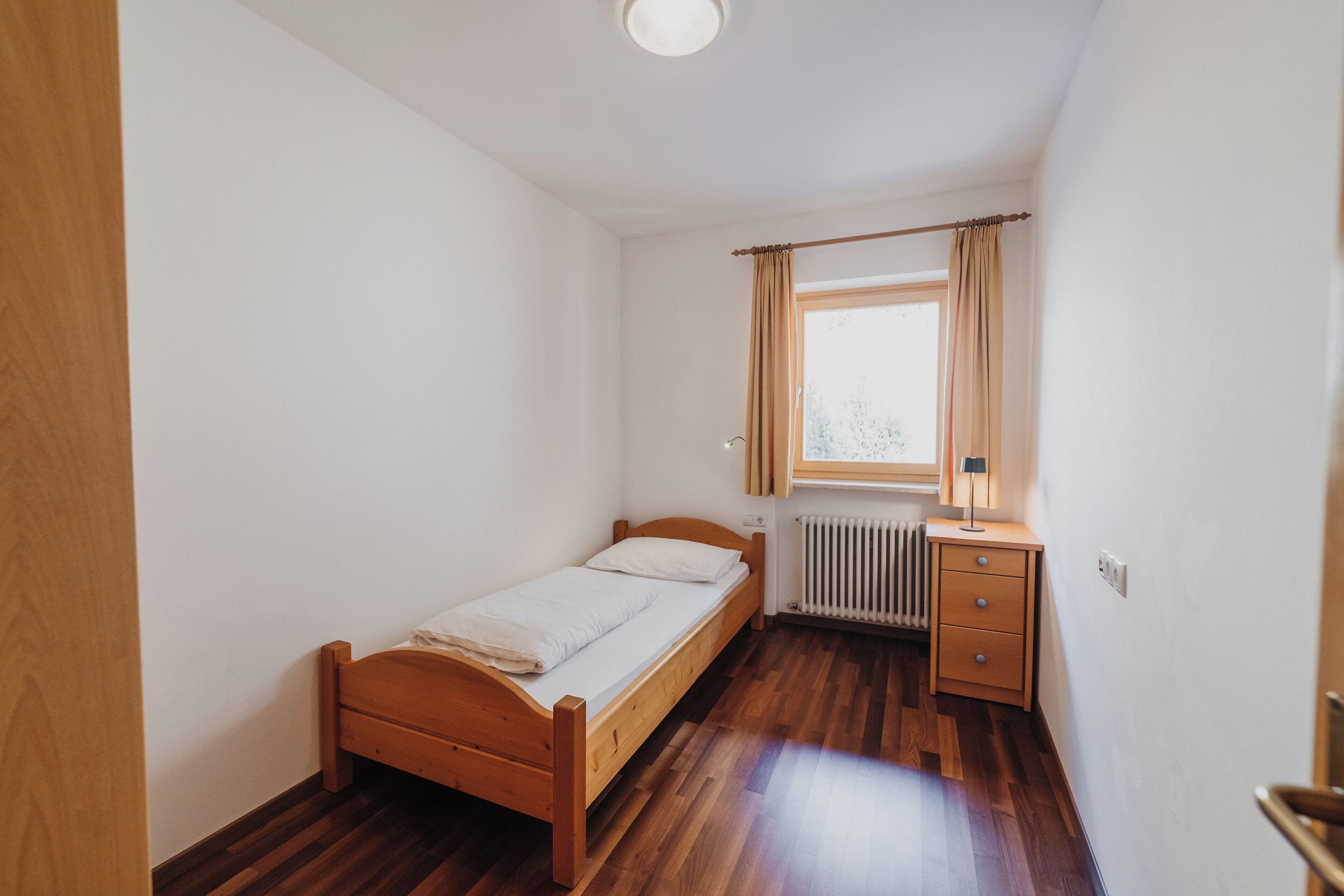 A small, clean room with a wooden bed, a desk with a lamp, and a radiator.
