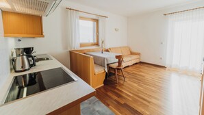A compact kitchen with a stove, sink, and coffee maker. There is a small dining area with a table and a chair. The room has wooden flooring and a window with curtains. - Appartements Elke (Lagundo)