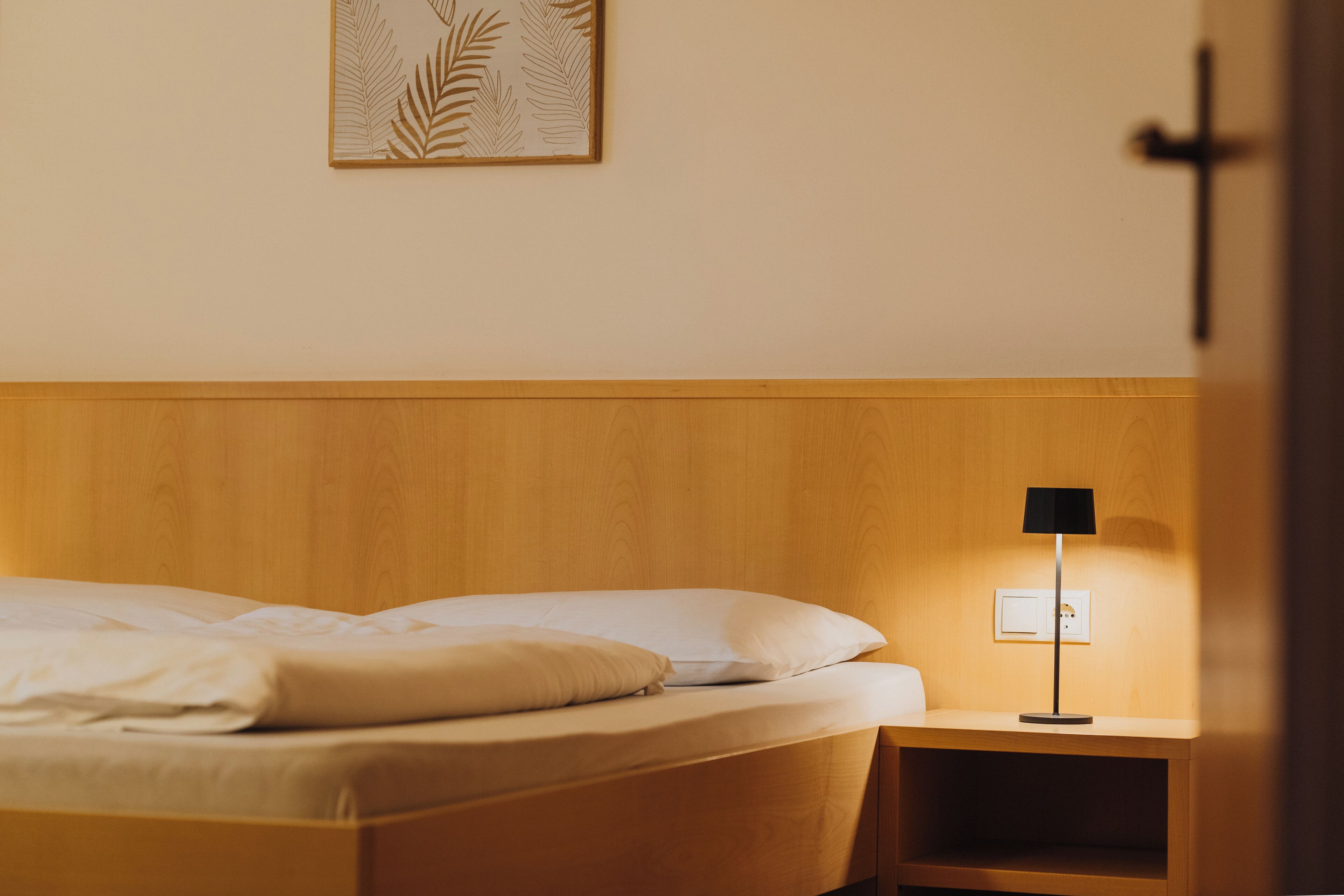 A neatly made bed with white linens, a bedside table with a lamp, and a wall-mounted power outlet.
