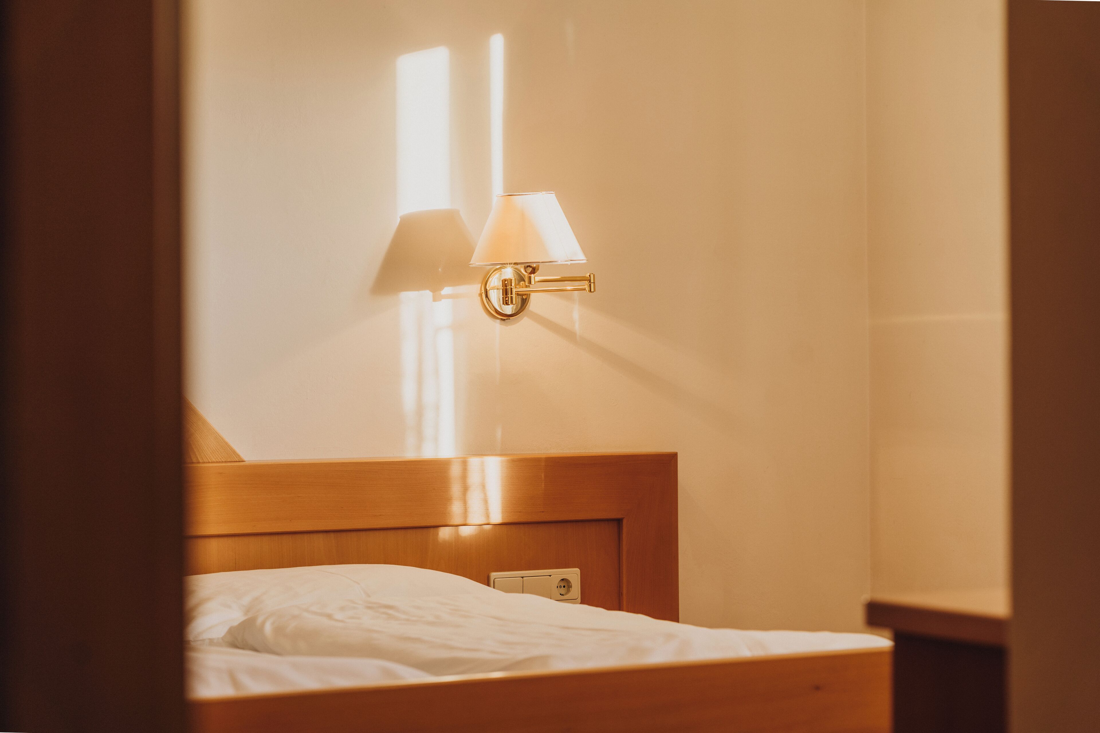 Una habitación de hotel con cabecera de madera, una cama individual con ropa blanca y una lámpara fijada a la pared.