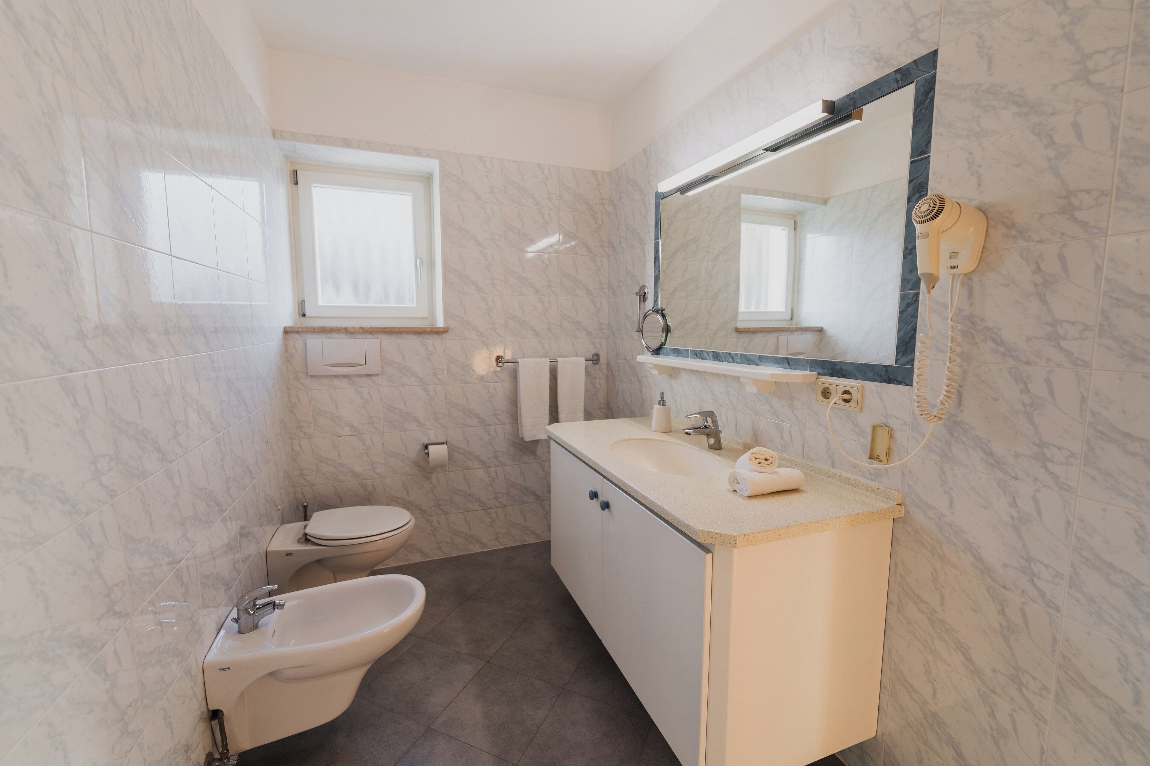 Une salle de bain équipée de toilettes, d’un bidet, d’un lavabo et d’un sèche-cheveux fixé au mur.
