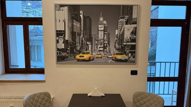 A dining area with a table and two chairs, a black and white cityscape painting on the wall, and large windows with a view of buildings.