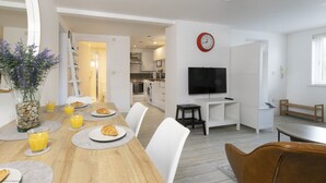 A modern kitchen with white cabinets, a built-in oven, and a microwave. There is a dining area with a wooden table, white chairs, and a vase with flowers. A TV is mounted on the wall, and a clock is visible.