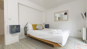 A modern bedroom with a wooden bed, white bedding, a bedside table with a lamp, a small cabinet, and a mirror on the wall.