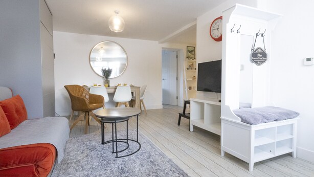 A modern living room with a sofa, a round mirror, a dining table with chairs, a TV, and a white cabinet.