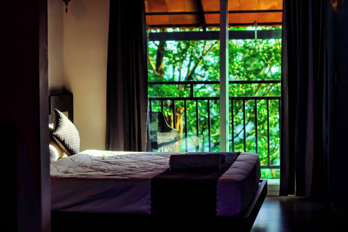 A bedroom with a bed, a nightstand, and a window with a view of greenery.