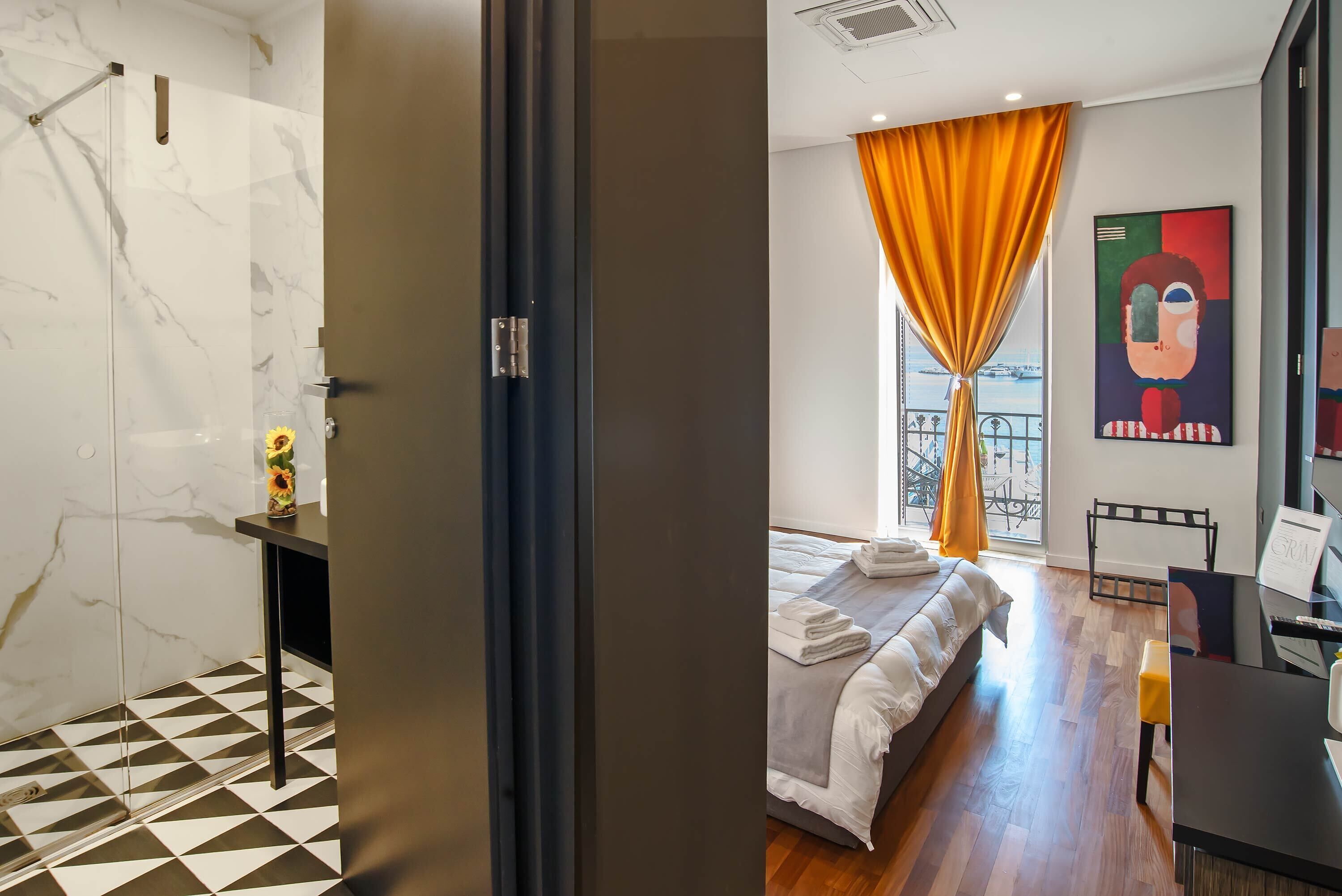 A modern hotel room with a shower, a bed with neatly folded towels, a desk, and a large window with orange curtains.