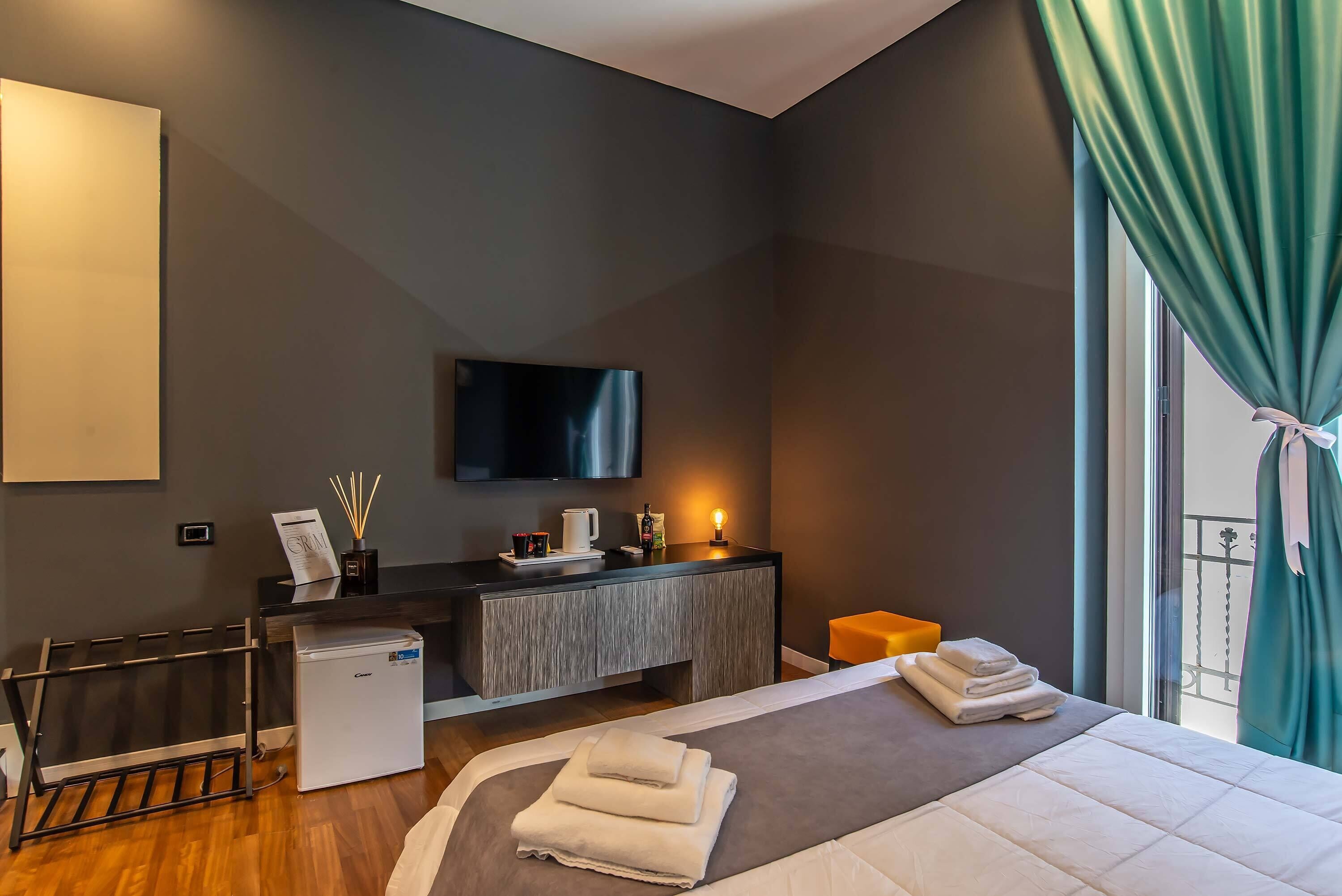 A modern hotel room with a bed, a small desk, a television, and a balcony with curtains.