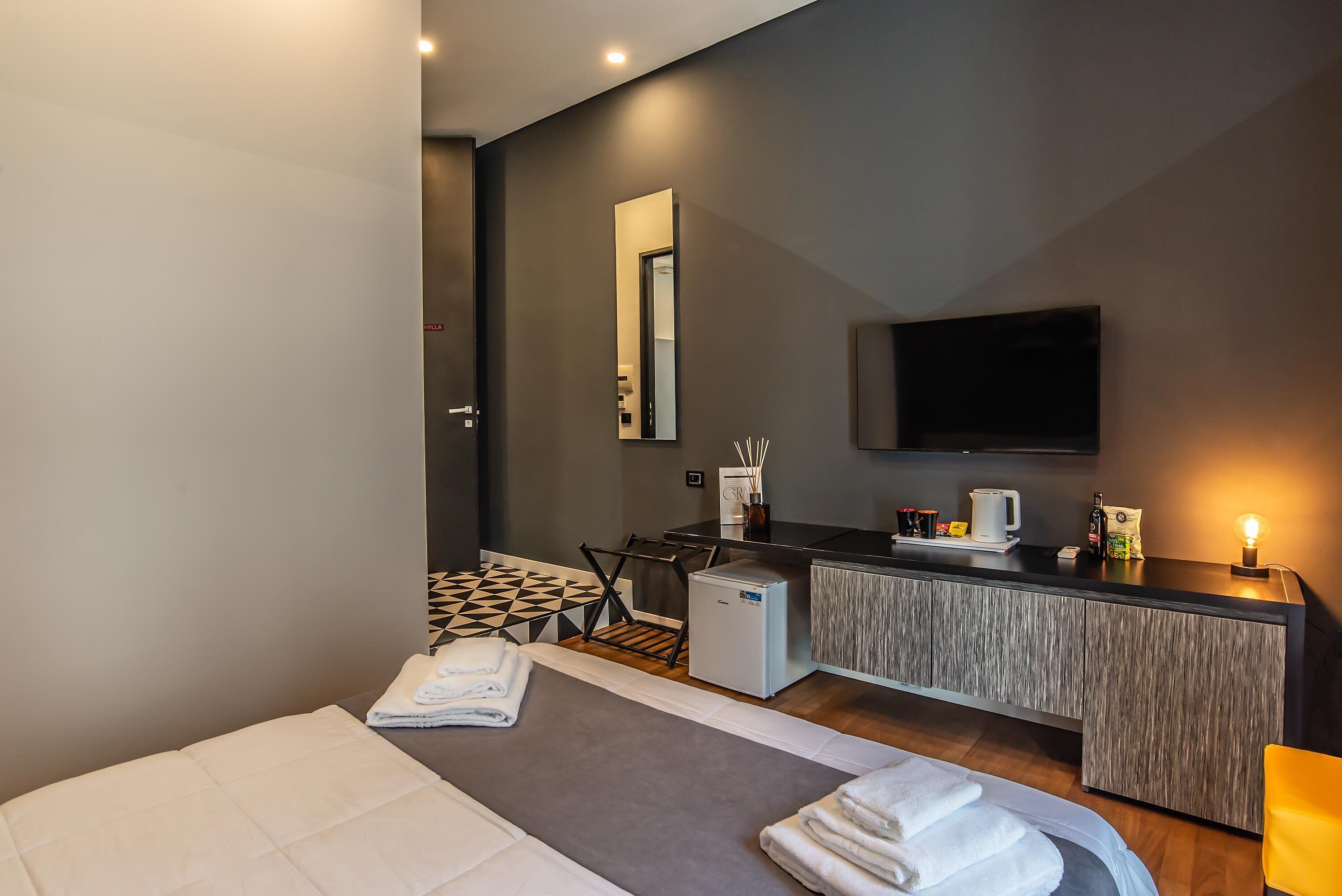 Une chambre d’hôtel moderne équipée d’un lit, d’un petit bureau avec une machine à café, d’un mini-réfrigérateur et d’un téléviseur à écran plat fixé au mur.