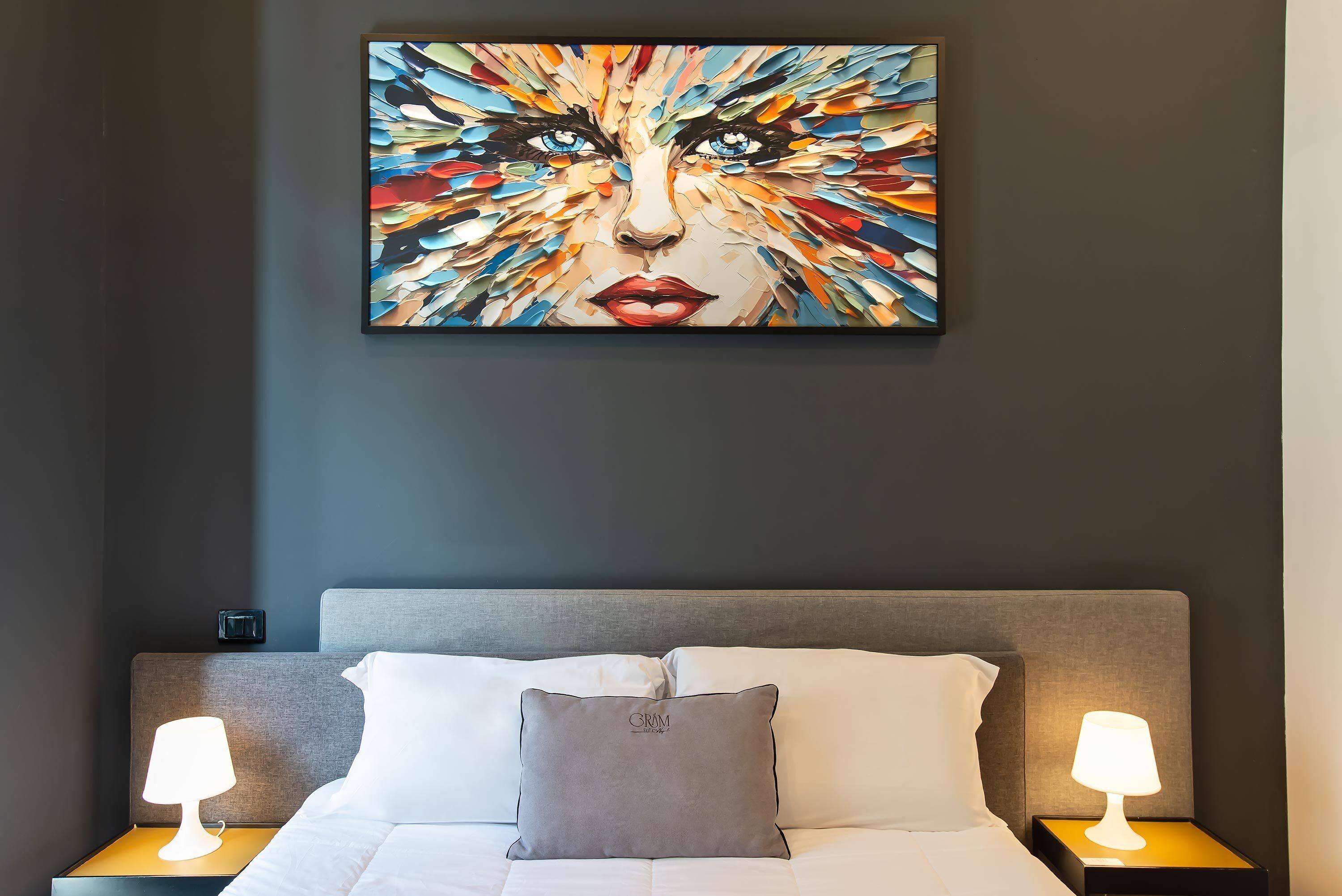 A hotel room with a large abstract painting on the wall, a bed with white bedding, a grey headboard, and two bedside lamps.