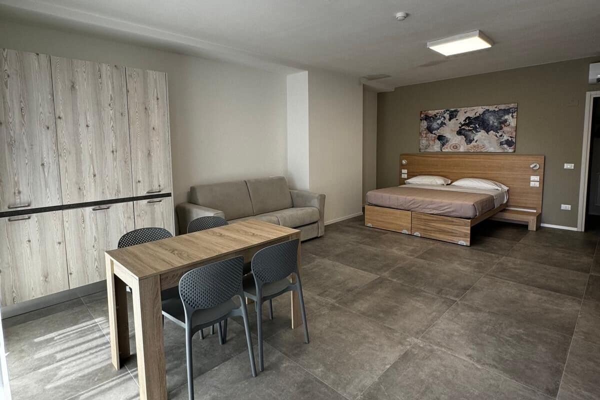 Une pièce moderne et bien éclairée, avec une table à manger en bois, deux chaises, un canapé et un lit doté d’une tête de lit en bois.