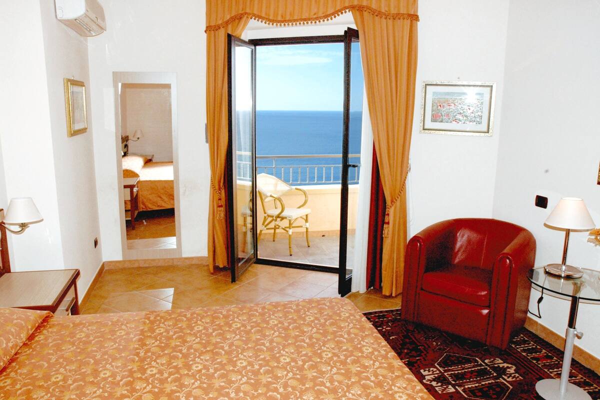 A hotel room with a bed, a red armchair, a glass table with a lamp, and a view of the sea through an open door.