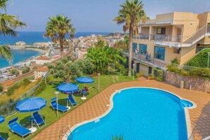 A swimming pool with blue umbrellas and lounge chairs, surrounded by palm trees and a view of the sea and town. - Nettuno Hotel Palace (Belvedere Marittimo)