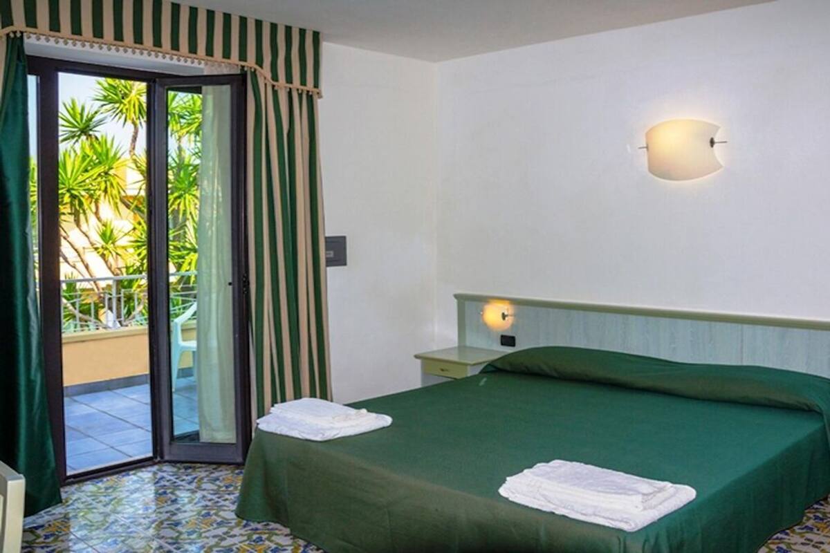 A hotel room with a bed, a nightstand, a lamp, a sliding glass door, and a view of palm trees.