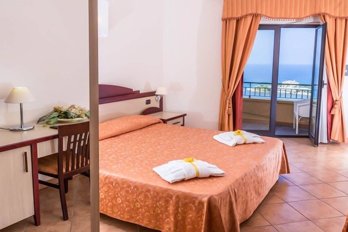 A hotel room with a bed, a desk, a chair, a nightstand, and a balcony with a view.