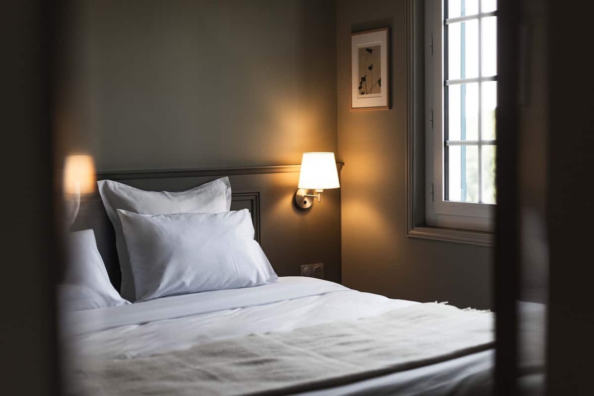 A neatly made bed with white linens, a wall-mounted lamp, and a framed picture on the wall.
