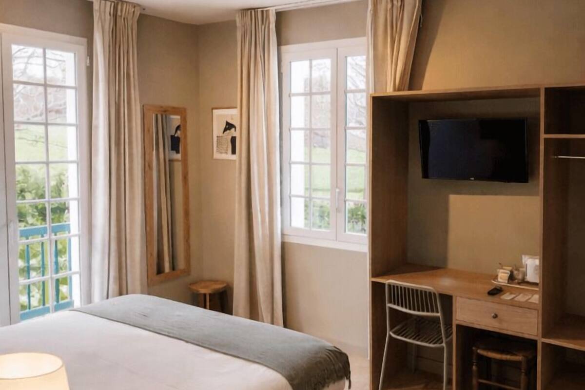 A hotel room with a bed, a desk, a chair, a TV, and large windows.
