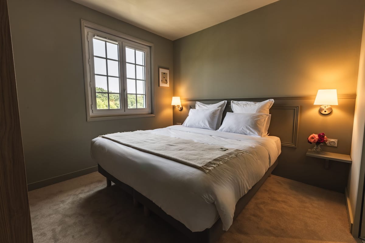 A neatly made bed with white linens and a beige throw, a bedside table with a lamp, and a window with a view of greenery.