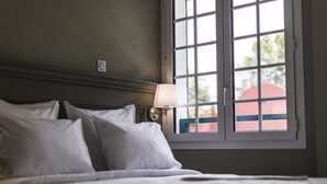 Ein ordentlich gemachtes Bett mit weißen Leinen und Kissen, eine Nachttischlampe und ein Fenster mit Blick auf ein Gebäude.