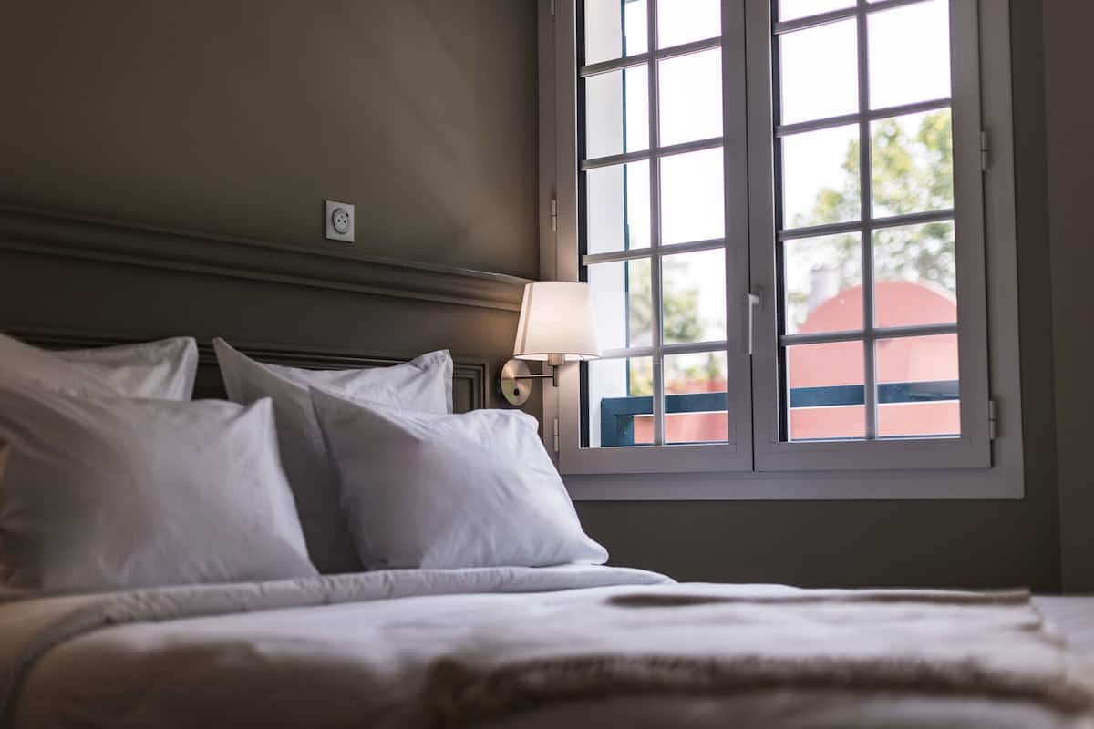 A neatly made bed with white linens and pillows, a bedside lamp, and a window with a view of a building outside.