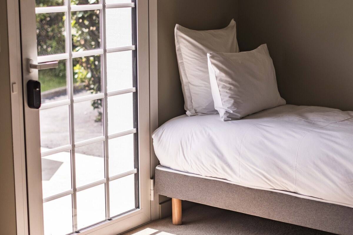 A bedroom with a bed, a door with a view of greenery outside, and a small cabinet above the bed.