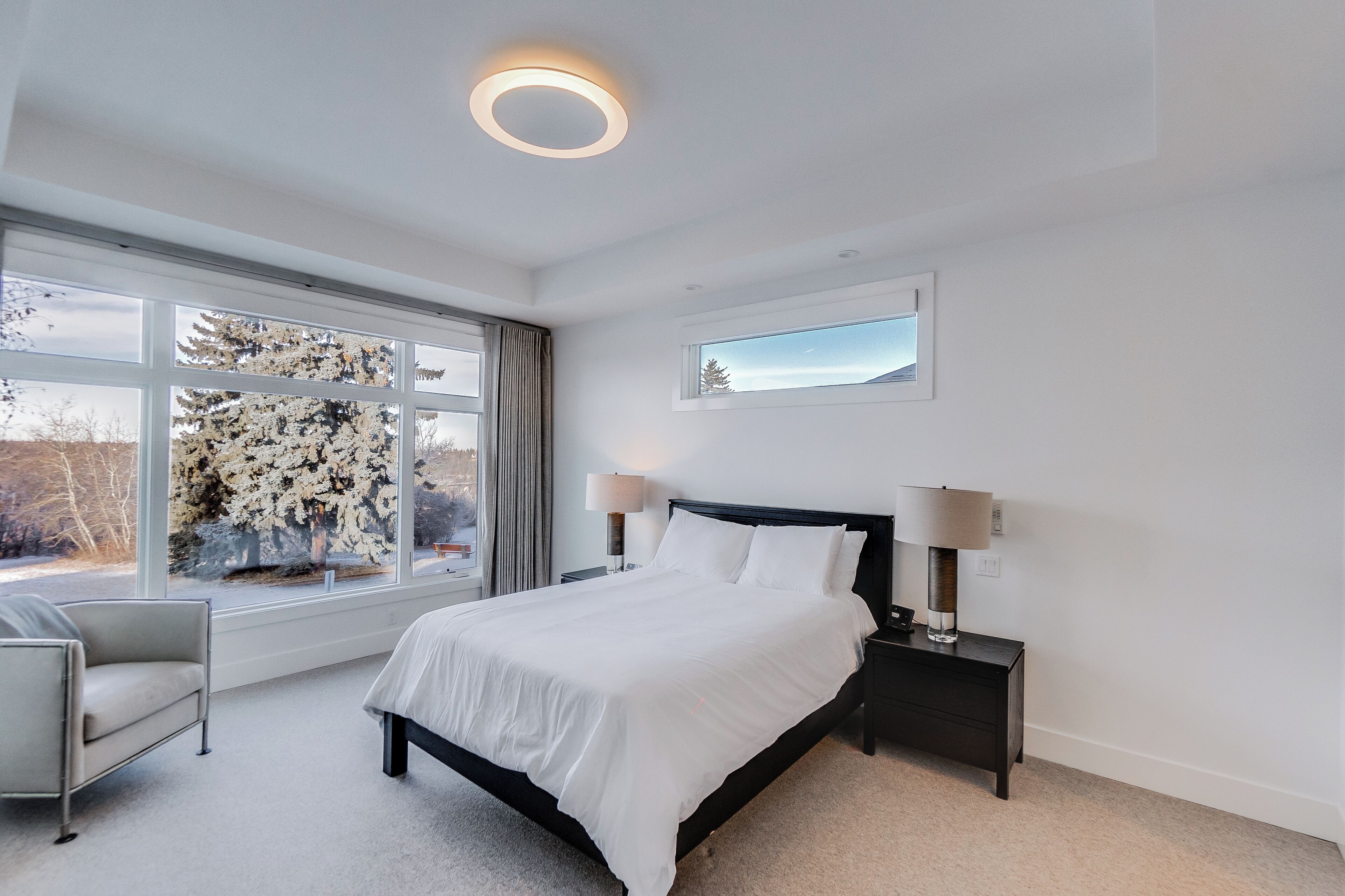 Ein modernes Schlafzimmer mit einem großen Bett, Nachttischlampen, einem Beistelltisch, einem Sessel und einem Fenster mit Blick auf Bäume.