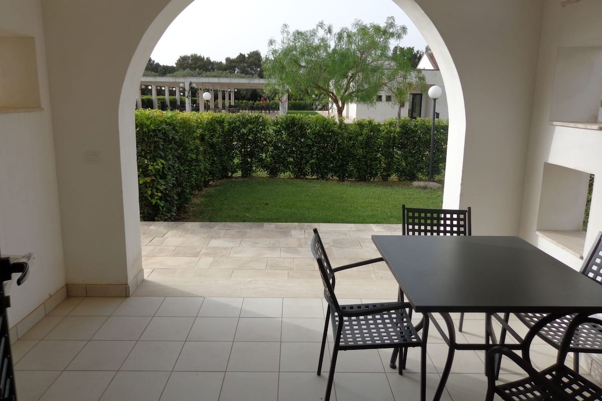 An outdoor patio area with a table and chairs, a tiled floor, and a view of a well-maintained garden through an archway.