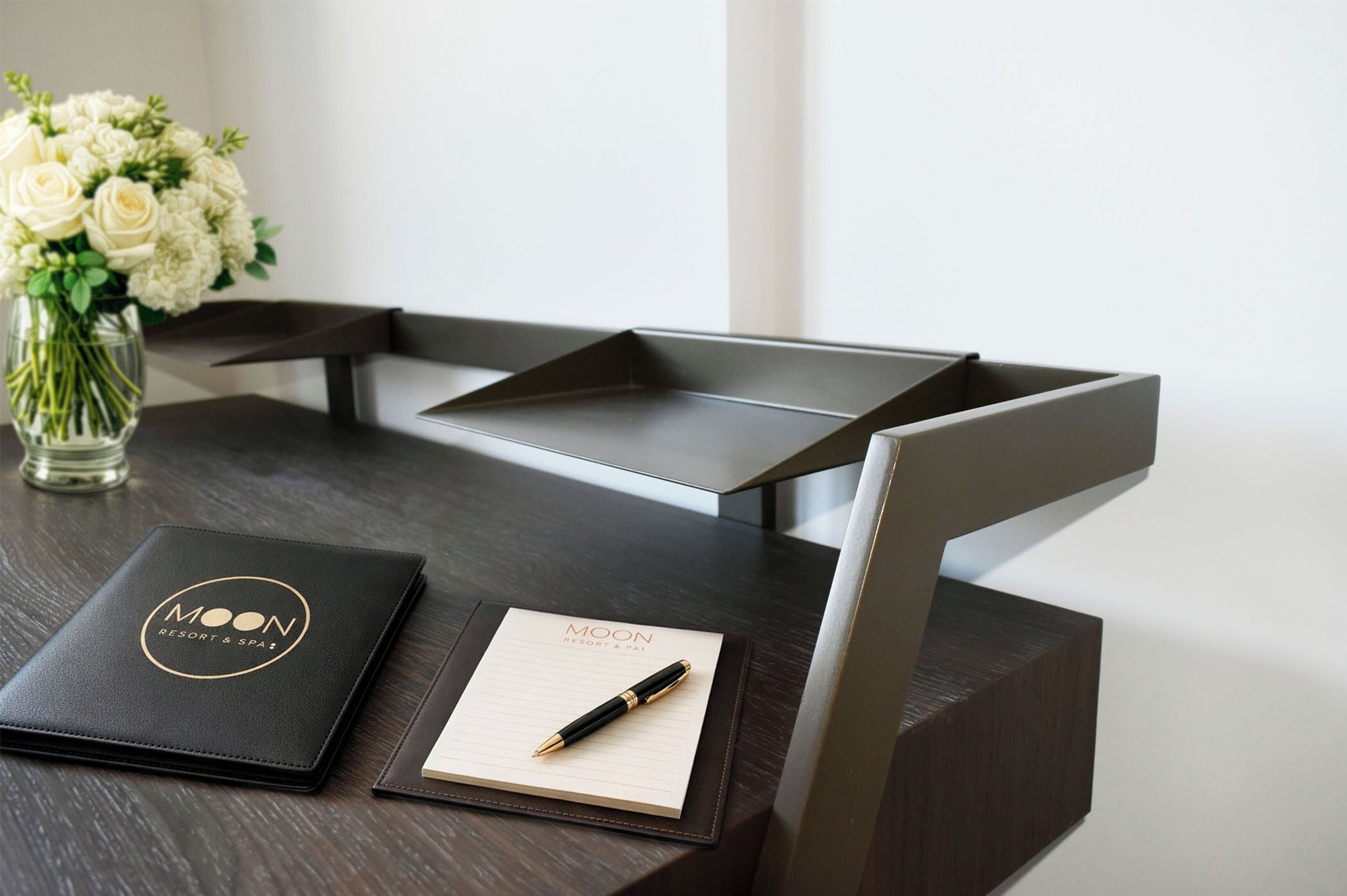 A desk with a black leather-bound notebook, a pen, and a glass vase with white flowers.