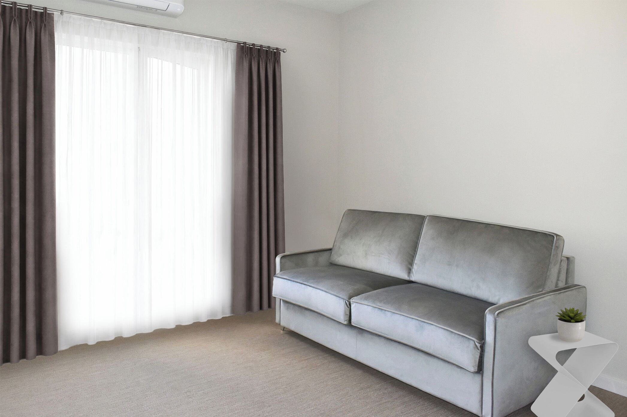 A modern living room with a grey sofa, a small white side table with a plant, and sheer curtains on a window.