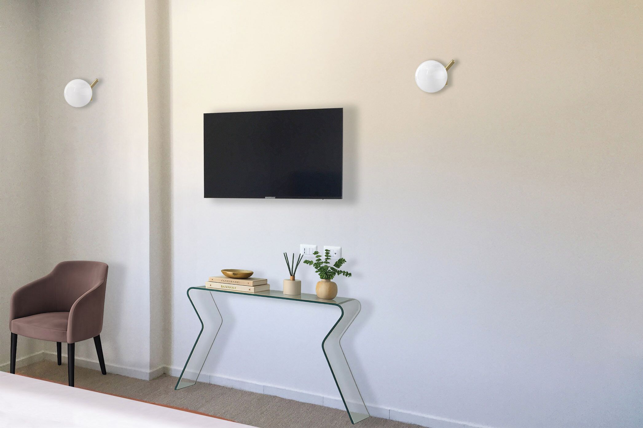 Une chambre moderne avec un téléviseur à écran plat fixé au mur, une console en verre agrémentée d’objets décoratifs, un fauteuil marron et une petite plante en pot.