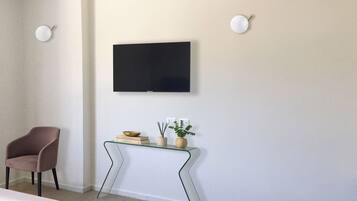 Une chambre moderne avec un téléviseur à écran plat fixé au mur, une console en verre agrémentée d’objets décoratifs, un fauteuil marron et une petite plante en pot.