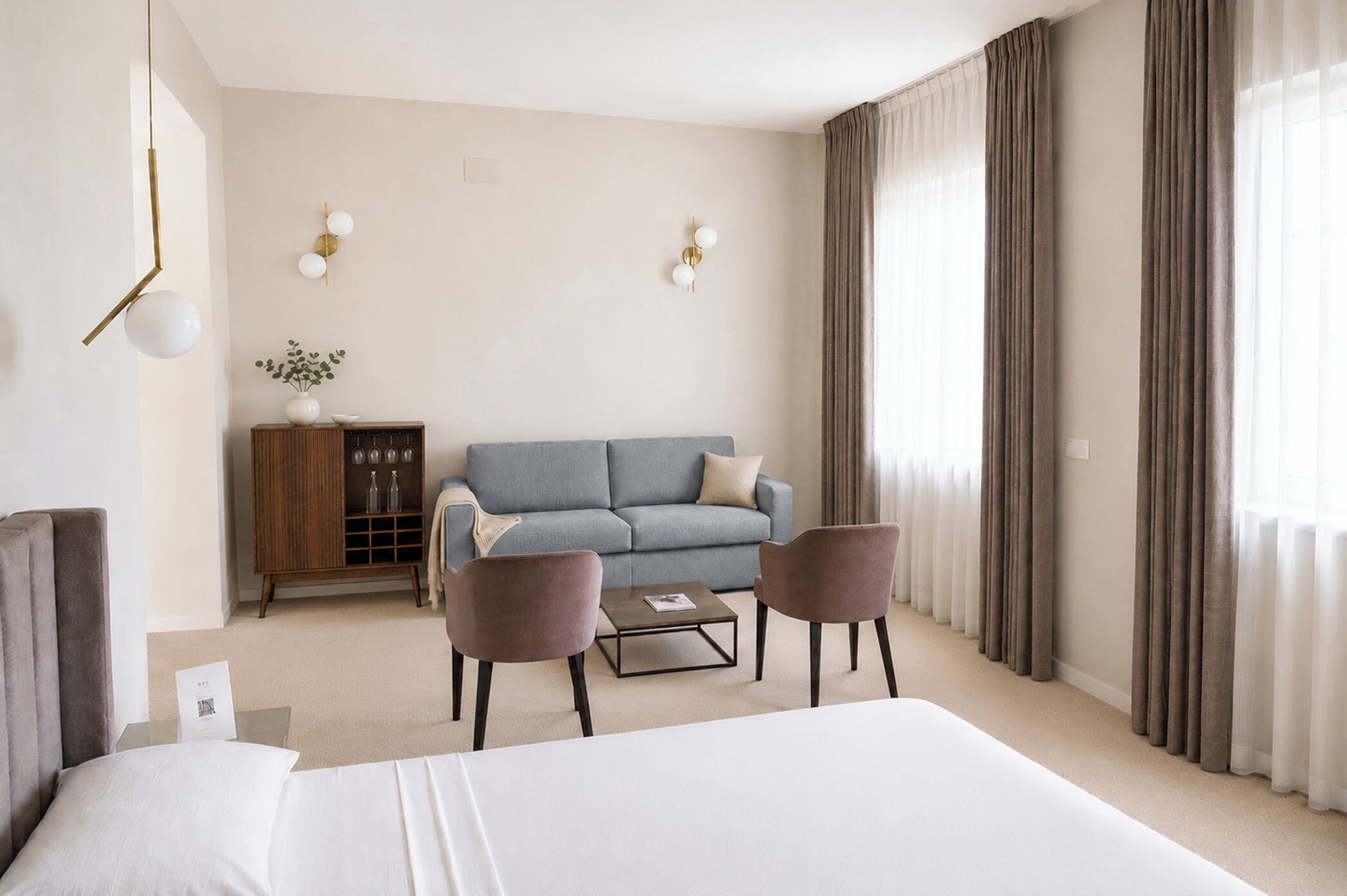 Ein Hotelzimmer mit einem Bett, einer Couch, zwei Sesseln, einem Couchtisch, einem Schrank mit Weingläsern und Vorhängen.