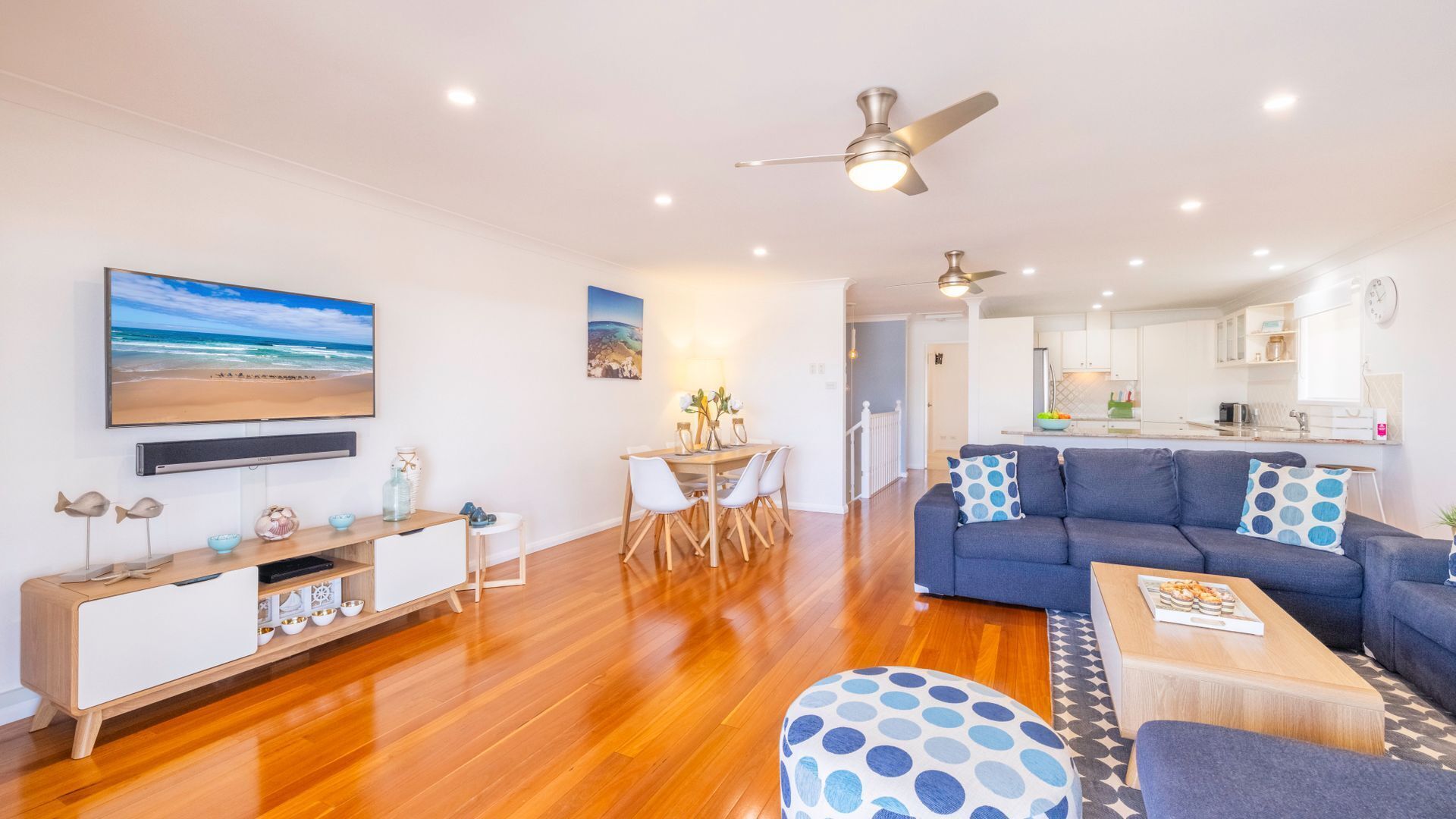 A modern living room with a wooden floor, a blue sofa, a dining area with a table and chairs, a kitchen in the background, and a ceiling fan.